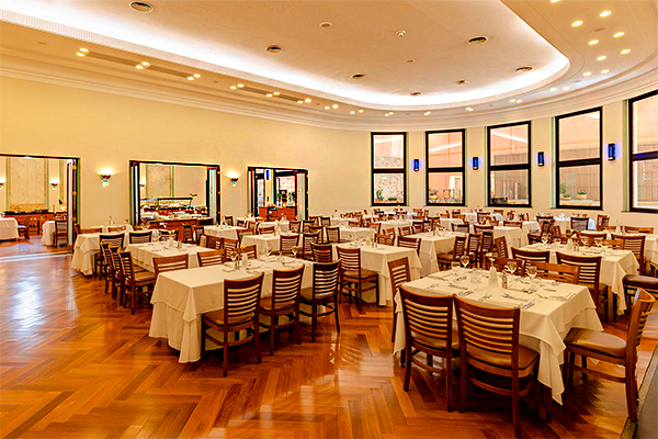 Dicas de Iluminação para Hotéis A imagem mostra o interior de um grande salão de restaurante ou refeitório. Há muitas mesas quadradas e retangulares, cobertas com toalhas brancas e com cadeiras de madeira, dispostas em fileiras no piso de madeira lustrosa. O salão tem uma forma levemente curva e é bem iluminado, com janelas altas e luzes no teto. No fundo à esquerda, portas abertas dão para outra área com um buffet.