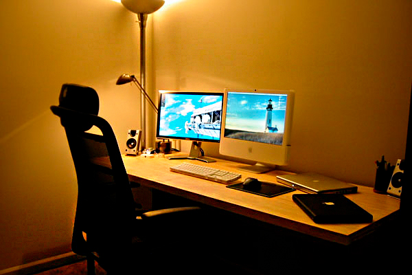 Como Iluminar o Home Office? A imagem mostra um home office com iluminação suave. Há uma mesa de madeira com dois monitores ligados, teclado, mouse, notebook e cadernos. Uma cadeira preta com encosto alto está à frente. No canto, uma luminária de coluna e uma de mesa iluminam o ambiente.