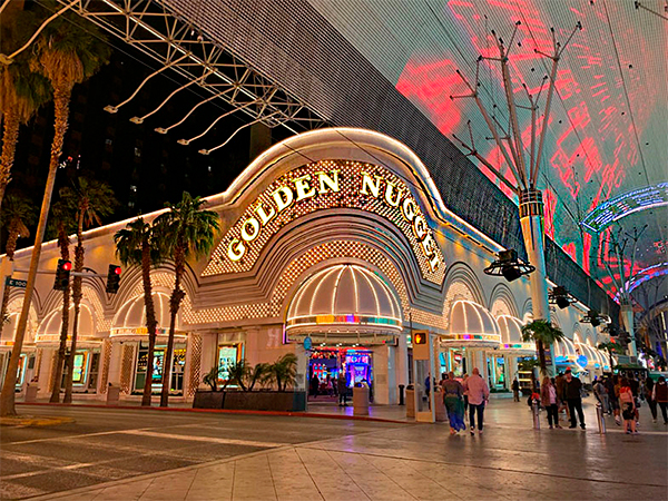Dicas de Iluminação para Hotéis Fachada iluminada do hotel e cassino Golden Nugget à noite, com letreiro dourado brilhante e luzes decorativas. Algumas pessoas caminham na calçada, cercadas por palmeiras e um teto coberto de luzes coloridas.