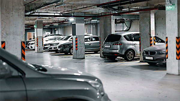 Dicas de Iluminação para Hotéis A imagem mostra uma garagem subterrânea iluminada, com colunas de concreto e várias vagas ocupadas por carros de diferentes modelos e cores. As colunas têm faixas laranja e pretas para sinalizar e proteger os veículos ao manobrar