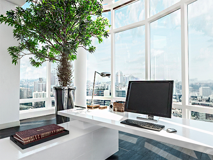 Como Iluminar o Home Office? A imagem mostra um escritório moderno e iluminado por luz natural. Há uma grande janela com vista para a cidade, uma mesa branca com computador, teclado, mouse e luminária metálica. Um vaso grande com uma árvore verde dá vida ao ambiente elegante e minimalista.