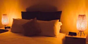 A imagem mostra um quarto escuro, provavelmente à noite. Há uma cama com travesseiros brancos e almofadas escuras. Em cada criado-mudo lateral, há um abajur aceso. As lâmpadas emitem uma luz amarelada e suave, criando uma atmosfera acolhedora para o sono.
