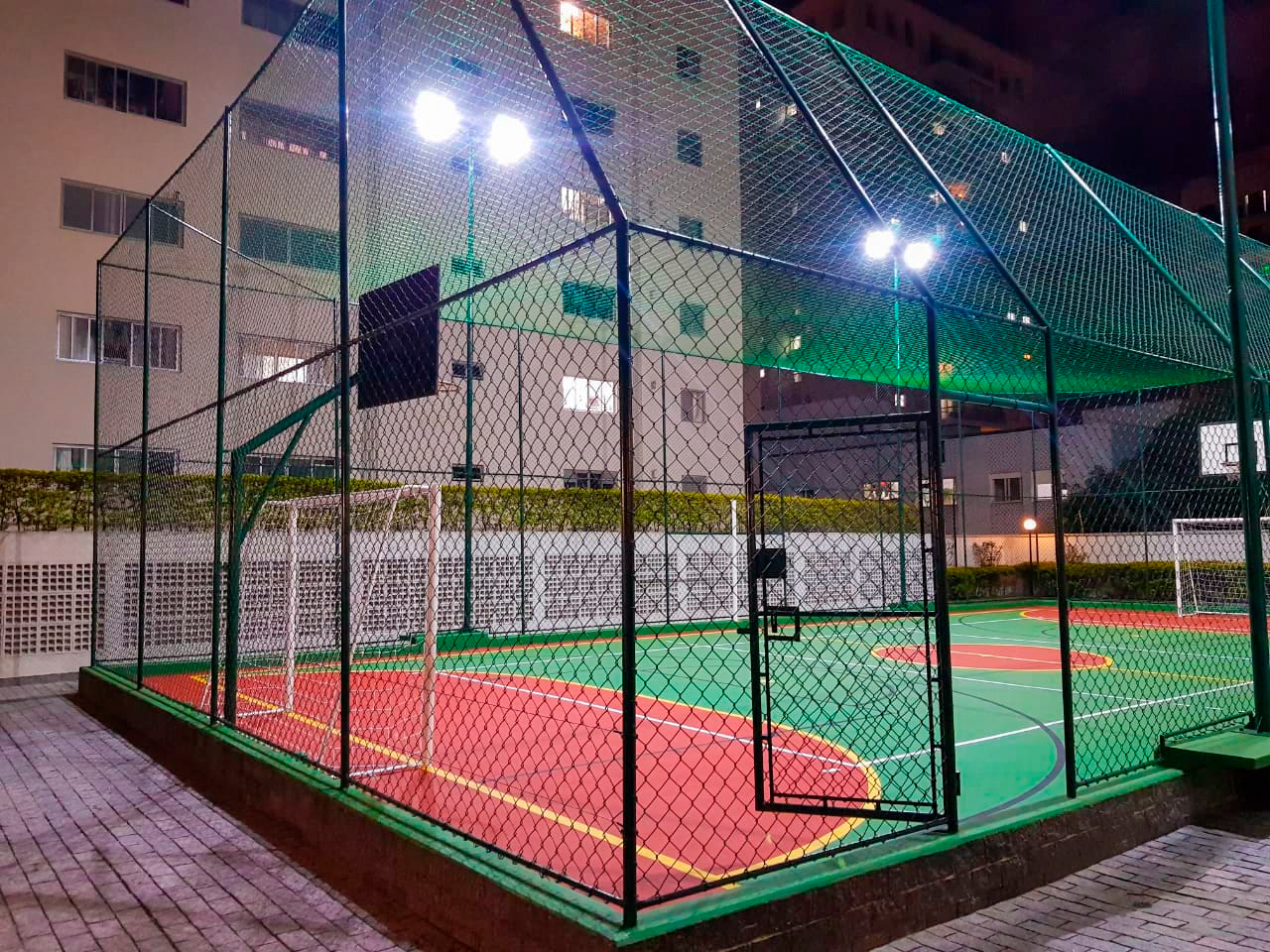 Dicas de Iluminação para Condomínios
A imagem noturna mostra uma quadra poliesportiva iluminada, cercada por uma tela de arame verde. O piso da quadra é dividido em áreas vermelhas e verdes, com marcações em amarelo. Há dois gols e cestas de basquete nas extremidades. Ao fundo, um prédio residencial se eleva