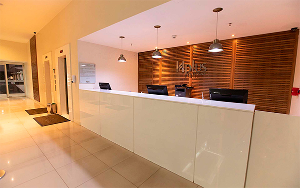 Dicas de Iluminação para Hotéis A imagem mostra a recepção de um hotel moderno. Há um balcão branco comprido com três computadores e luminárias pendentes. Ao fundo, uma parede de madeira com o nome do hotel em metal. O ambiente é bem iluminado e limpo, com piso claro e decoração minimalista.