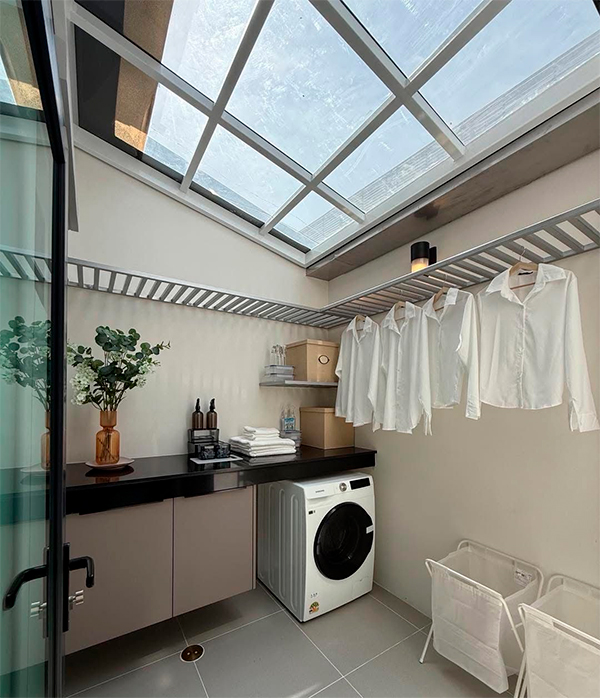 Projete uma Iluminação para Sua Lavanderia Uma bancada escura sobre armários cor de areia/bege. Uma máquina de lavar roupa branca no centro. Prateleiras de metal elevadas com cabides onde camisas brancas estão penduradas. Cestos de roupa branca no chão. Piso de azulejo cinza claro.